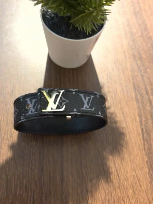 Armband  - Snyggt svart armband med klassiskt -monogram i grått och silverfärgat -spänne framtill. Tillverkat i ett tåligt material med exklusiv känsla och stilren design. Perfekt accessoar för dig som gillar lyxiga detaljer och vill sticka ut.