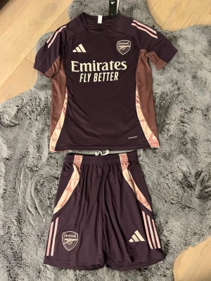 Arsenal shortset i vinrött - Snyggt Arsenal shorts träningsset från Adidas i vinrött med rosa detaljer. Setet består av en kortärmad tröja och matchande shorts, båda med klubbmärke och Adidas-logga. Materialet är lätt och andas, perfekt för fotbollsträning eller gymmet. Storlek S men den passar M perfekt