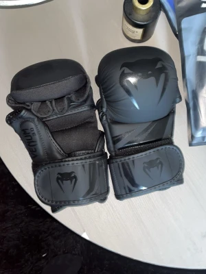 Svarta MMA-handskar från Venum - Säljer ett par svarta MMA-handskar från Venum med coolt ormlogga på ovansidan. Handskarna har vaddering för extra skydd och kardborreband runt handleden för bra passform. Perfekta för träning inom kampsport eller sparring.