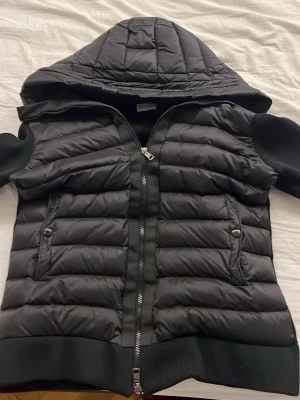 Svart dunjacka med huva Moncler - Snygg svart dunjacka från Moncler med quiltat mönster, huva och ribbade muddar. Jackan har dragkedja framtill, två fickor med knappstängning och Moncler-logga på ärmen. Perfekt för kalla vinterdagar och höstdagar och riktigt stilren look. Använd 3/4 gånger 