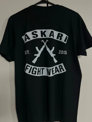 Svart Askari Fight Wear t-shirt - Svart t-shirt med stort vitt tryck på ryggen från Askari Fight Wear. Trycket har två korsade gevär, texten 'ASKARI FIGHT WEAR' och 'EST. 2015'. Klassisk passform och rund hals. Perfekt för dig som gillar streetwear och kampsport.