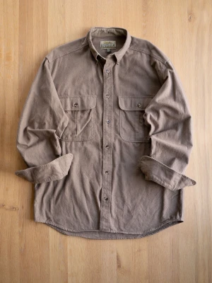 Vintage Beige Flanell - Säljer en beige overshirt från Cabela's i mjukt material. Notera skadan på armbågen! Skjortan har två stora bröstfickor med lock och knappar framtill. Perfekt för lager-på-lager och har en avslappnad passform med långa ärmar.
