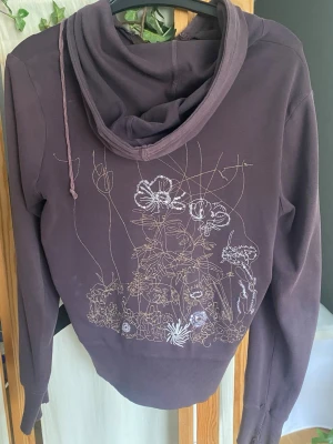 Lila hoodie med blommig brodyr - Säljer en lila hoodie med dragkedja och huva. På ryggen finns en detaljerad brodyr med blommor och växter i vitt och lila. Hoodien har långa ärmar, ribbade muddar och en avslappnad passform. Perfekt för dig som gillar unika detaljer och skön stil. Står L men passar snarare som s/m