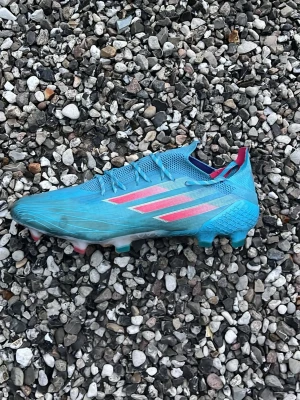 Blåa Adidas fotbollsskor - Säljer ett par blåa Adidas fotbollsskor med rosa och vita detaljer på sidorna. Skorna har låg profil, snörning och dobbar för spel på gräs. Tillverkade i syntetmaterial för lätt vikt och bra bollkontroll. Perfekta för dig som vill sticka ut på planen.