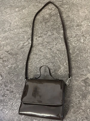 Mörkbrun lackad axelväska - Snygg mörkbrun axelväska i lackat material med fyrkantig form och lång axelrem. Väskan har ett litet handtag upptill och stängs med lock. Perfekt för dig som gillar en stilren och trendig look.