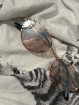 Ovala solglasögon med roséguld båge - Snygga solglasögon med tunn roséguldfärgad metallbåge och ovala, lätt tonade linser. Skalmarna är svarta och näskuddarna är transparenta för extra komfort. Perfekt accessoar för att lyfta din stil i solen.