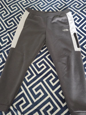 Gråa mjukisbyxor från The North Face - Snygga gråa mjukisbyxor från The North Face med vita paneler längs sidorna och en svart dragkedjeficka på höger ben. Byxorna har resår i midjan och är tillverkade i ett mjukt och bekvämt material, perfekta för chill dagar.