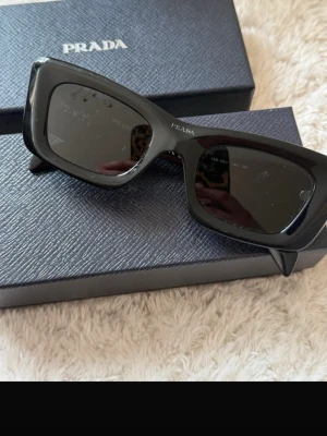 Svarta solglasögon från Prada - Snygga svarta solglasögon från Prada med rektangulära bågar och diskret logga på sidan. Glasögonen har en stilren och modern design som ger en cool vibe till din look. Perfekta för dig som vill sticka ut med en lyxig accessoar.