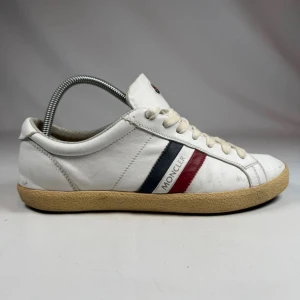 Vita Moncler sneakers  - Snygga vita sneakers från Moncler i läder med klassiska röd och blå ränder på sidan och logga på plösen. Skorna har vita snören och en beige gummisula som ger en retrovibe. Perfekta för dig som gillar stilrena och sportiga skor.