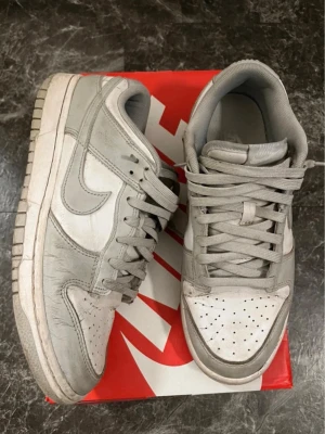 Nike Dunk Low gråvita sneakers - Nike Dunk Low sneakers i grått och vitt med klassisk låg siluett. Skorna har perforerad tå, vita paneler och gråa detaljer samt matchande gråa skosnören. Tillverkade i läder med gummisula och ikonisk Swoosh på sidan.