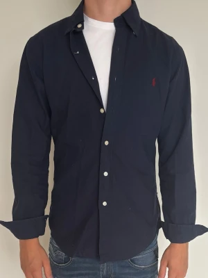 Mörkblå ralph luaren skjorta  - Klassisk mörkblå ralph luaren skjorta med långa ärmar och vita knappar framtill. Skjortan har en liten röd broderad logga på bröstet och normal passform. Perfekt till jeans eller chinos för en clean look.