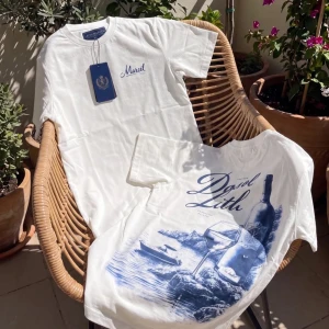 Vit t-shirt med blått tryck - Säljer en vit t-shirt med blått tryck och texten 'Mariel' på bröstet. Baksidan har ett stort konstnärligt motiv i blått med natur och flaskdetaljer. Klassisk passform och rund hals. Perfekt för dig som gillar unik design och stilrena färger.