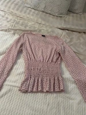 Ljusrosa prickig blus med volang - Supersöt ljusrosa blus med svarta prickar och smockad midja som ger en snygg siluett. Långa ärmar och volang nedtill för extra detalj. Perfekt att styla med jeans eller kjol för en trendig look.