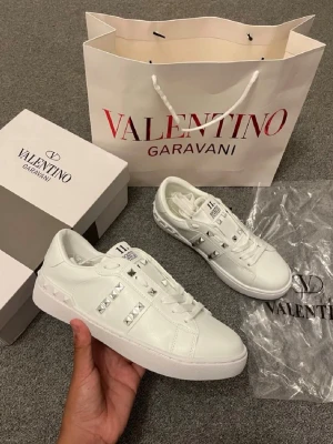 Vita Valentino Garavani sneakers - Säljer ett par vita sneakers från Valentino Garavani med coola silverfärgade nitar på sidorna. Skorna är i läder och har en klassisk låg siluett med vita snören och diskret logga på plösen. Perfekta för dig som vill sticka ut med stilrena detaljer.
