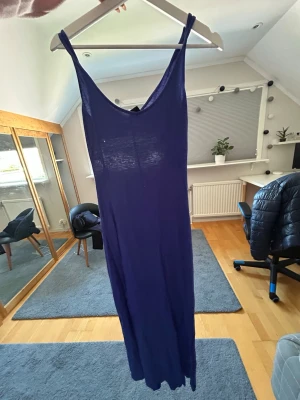 Lång blå ärmlös klänning - En enkel och stilren lång blå klänning med smala axelband och djup urringning både fram och bak. Klänningen är ärmlös och har en rak passform, perfekt för varma dagar. Materialet ser ut att vara tunt och mjukt, troligen bomull eller viskos.