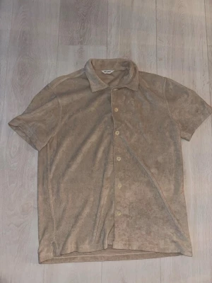 Beige frottéskjorta från East West - Säljer en beige skjorta från East West i mjuk frotté. Skjortan har korta ärmar, klassisk krage och knappar framtill. Perfekt för en chill och avslappnad stil. Passar dig som gillar unika material och vill ha något annorlunda i garderoben.