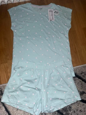 Mintgrön pyjamas med hjärtan  - Tvådelad pyjamas från Pepco i mintgrönt med små vita hjärtan. Setet består av en kortärmad t-shirt och shorts med resår i midjan. Perfekt för mysiga kvällar hemma. Materialet är mjukt och bekvämt.