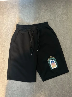 Svarta shorts från Casablanca - Svarta shorts från Casablanca med broderad detalj på ena benet och texten 'Avenida Casablanca'. Shortsen har resår i midjan med snörning och sidofickor. Perfekta för en avslappnad stil.