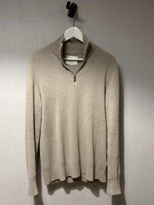 Beige ribbstickad tröja med halvzip - Snygg beige ribbstickad tröja med halvzip och hög krage. Tröjan har långa ärmar och är tillverkad i ett mjukt, stretchigt material som ger en avslappnad men stilren look. Perfekt för lager på lager under kyliga dagar.