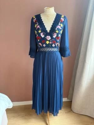 Blå långklänning med broderade blommor asos design stl 36 - Superfin blå långklänning med v-ringning och broderade blommor i rött, rosa och gult. Klänningen har trekvartsärmar med spetsdetaljer och plisserad kjol. Perfekt för dig som vill sticka ut med färg och detaljer! Klänningen är som ny. 