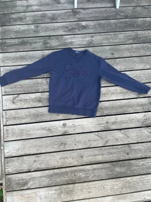 Blå sweatshirt från Polo Ralph Lauren - Snygg blå sweatshirt från Polo Ralph Lauren med broderad logga på bröstet. Tröjan har rund halsringning och ribbade muddar vid ärmslut och nederkant. Perfekt för en avslappnad och stilren look.