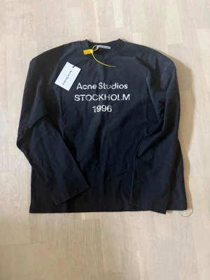 Svart långärmad topp Acne Studios - Svart långärmad topp från Acne Studios med tryck 'Acne Studios STOCKHOLM 1996' i vitt framtill. Klassisk rund halsringning och rak passform. Perfekt för dig som gillar stilrena och trendiga plagg. DEN ÄR BAGGY I STORLEKEN