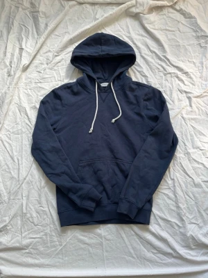 Mörkblå hoodie från Pier One - Mörkblå hoodie från Pier One med klassisk känguruficka och vita dragsnören i huvan. Tillverkad i mjukt och bekvämt material, perfekt för en avslappnad stil. Långa ärmar och ribbade muddar vid ärmslut och nederkant.