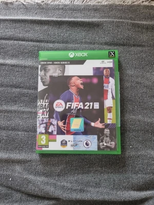 FIFA 21 till Xbox One/Series X - FIFA 21 är ett fotbollsspel till Xbox One och Xbox Series X med omslag i grönt plastfodral. Spelet har EA Sports-logga och visar Kylian Mbappé på framsidan. Omslaget har inslag av svart, vitt, blått och rött samt flera fotbollsbilder.