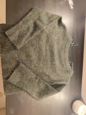 Gråmelerad stickad tröja - Mysig grön stickad tröja med rund halsringning och långa ärmar. Tröjan har en fluffig, lite luddig yta och ribbstickade muddar vid ärmslut och nederkant. Perfekt för kyliga dagar och enkel att matcha med olika outfits.