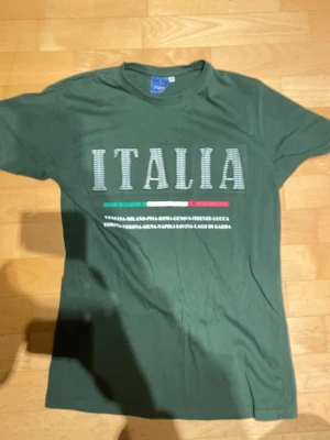 Grön t-shirt med ITALIA-tryck - Mörkgrön t-shirt från Tutto T-Shirt med stort ITALIA-tryck på bröstet och text i italienska färger. Klassisk rund hals och korta ärmar. Perfekt för dig som gillar enkel stil med en touch av Italien.