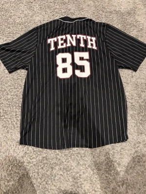 Svart randig baseballskjorta - Säljer en svart baseballinspirerad skjorta med vita tunna ränder och trycket 'TENTH 85' på ryggen. Skjortan har knappar framtill och korta ärmar. Perfekt för dig som gillar sportig stil och vill sticka ut lite extra.