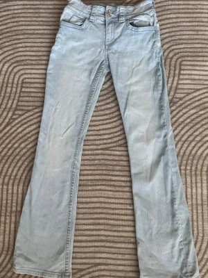 Nya Ljusblå bootcut jeans - Snygga ljusblå jeans med bootcut passform och klassiska fem fickor. Jeansen har dubbla knappar i midjan och detaljerade bakfickor med knappstängning. Perfekta för en avslappnad och trendig look.