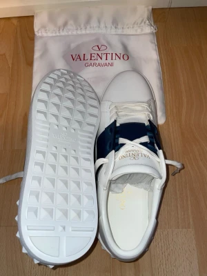 Valentino Garavani vita sneakers - Snygga vita sneakers från Valentino Garavani med en bred mörkblå rem över sidan. Skorna har vita snören, diskret logga på plösen och en unik sula med rutmönster. Tillverkade i läder och levereras med dustbag. SKRIV FÖR ATT DISKUTERA PRISET