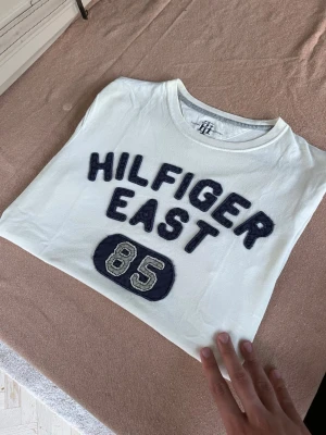 Vit Hilfiger East t-shirt med tryck - Riktigt fet Tommy hilfiger ”Hilfiger East 85” T-shirt med påsytt tryck! Storlek S. Hör vad dig vid frågor😁