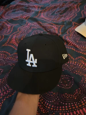 Svart LA New Era 59FIFTY keps - Säljer en svart New Era 59FIFTY keps med LA-logga broderad i vitt framtill och New Era-logga på sidan. Klassisk flat brim och strukturerad passform. Perfekt för dig som gillar streetwear och sportig stil. Aldrig använt.
