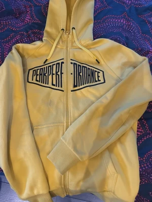 Gul peakperformanse hoodie med dragkedja  - Säljer en gul hoodie med dragkedja och svart tryck på bröstet där det står 'Peak Performance'. Hoodien har huva med snören och två fickor framtill. Perfekt för en avslappnad och sportig stil.