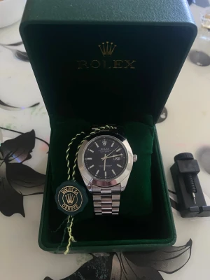 R*lex daydate - Säljer en stilren R*lex klocka med mörkblå urtavla och datumvisning. Klockan har ett klassiskt armband i silverfärgat stål och levereras i originalbox med tagg. Perfekt accessoar för dig som gillar exklusiv och tidlös design. Följer med ett accessoar som kan justera storleken av klockan till din arm.