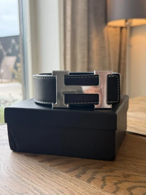 Svart bälte med stor silver H-spänne - Snyggt svart bälte i skinn med en stor, blank H-formad silverspänne. Bältet har vita kontrastsömmar längs kanterna som ger en cool detalj. Perfekt accessoar för att lyfta vilken outfit som helst.