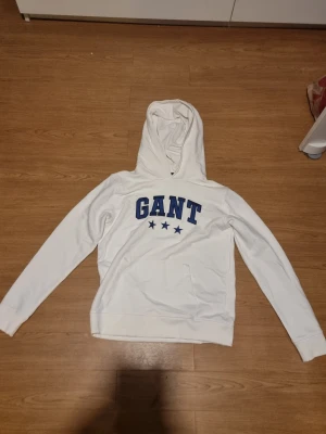 Vit hoodie från GANT med blå text - INSTABOX!!! Snygg vit hoodie från GANT med blå broderad logga och tre stjärnor på bröstet. Klassisk känguruficka framtill och ribbade muddar vid ärmslut och nederkant. Perfekt för en avslappnad och sportig stil.