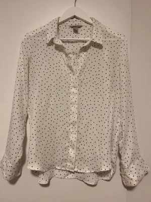 Vit blus med svarta prickar - Snygg vit blus med svarta prickar och klassisk krage. Blusen har långa ärmar och knäppning framtill. Tillverkad i ett lätt och luftigt material som ger en skön känsla. Perfekt att styla med jeans eller kjol för en trendig look.