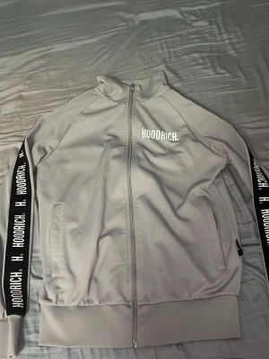 Grå zip hoodie från Hoodrich - Snygg grå zip hoodie från Hoodrich med vit logga på bröstet och svarta band med vit text längs ärmarna. Tillverkad i mjukt och stretchigt material, har två fickor framtill och ribbade muddar. Perfekt för en avslappnad streetwear-look.