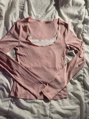 Ljusrosa ribbad långärmad topp - Säljer en ljusrosa ribbad topp med lång ärm och fyrkantig ringning. Toppen är figurnära och har en mjuk, stretchig känsla som passar perfekt till jeans eller kjol. Enkel men ändå snygg detalj med den breda ringningen framtill.