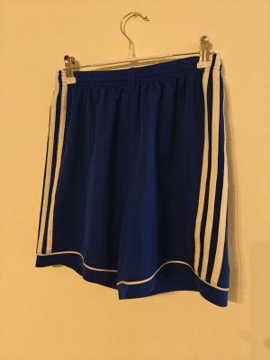 Adidas träningsshorts  - Snygga blå sportshorts från Adidas med klassiska vita ränder längs sidorna. Shortsen har elastisk midja och är tillverkade i ett lätt och svalt material som passar perfekt för träning eller sportiga outfits. Storlek L för barn.