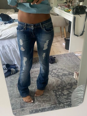 Blå slitna bootcut jeans - Säljer ett par blå bootcut jeans med slitningar och hål på benen. Jeansen har låg midja, klassiska fem fickor och ljusare tvättade partier. Perfekta för en avslappnad och trendig look.