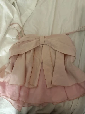 Ljusrosa volang topp med rosett - Supersöt ljusrosa topp med tunna axelband och stor rosett framtill. Toppen har volanger i flera lager och är tillverkad i ett lätt, luftigt material som ger en romantisk vibe. Perfekt för dig som vill ha en feminin och trendig look.