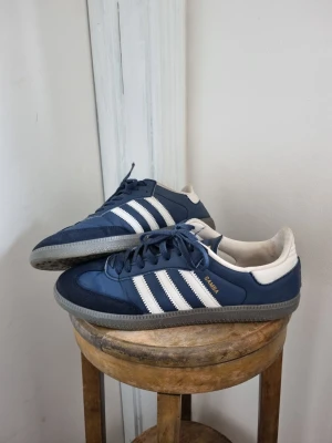 Adidas Samba blå och vita sneakers - Klassiska Adidas Samba sneakers i blått med vita detaljer. Ovandelen är i läder och mocka. Strl 43,5 men passar även 43 Släppt lite på ena skon som förmodligen går att limma och något litet hål under båda. 