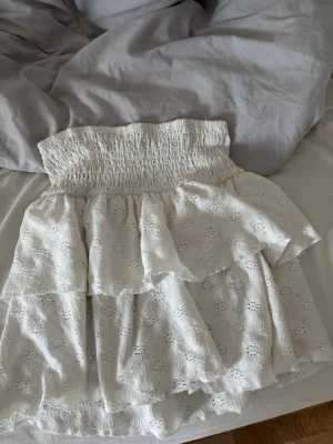Vit volangkjol med broderade detaljer - Supersöt vit kortkjol med bred smock i midjan och flera lager volanger. Kjolen har broderade blommönster och är gjord i ett lätt bomullstyg, perfekt för varma dagar. Passar dig som gillar romantisk och feminin stil.