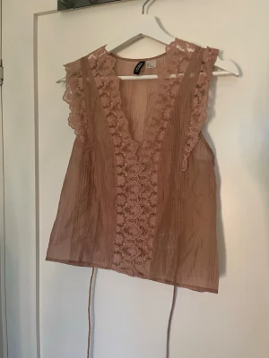 Beige spetsdetaljerad linnetopp Divided - Säljer en beige linnetopp från Divided med v-ringning och breda spetsdetaljer längs kanterna. Toppen är ärmlös och har tunna knytband i sidan. Perfekt för sommarens alla tillfällen och ger en romantisk vibe.