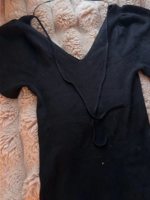 Svart ribbad klänning med snörning - Säljer en svart ribbad klänning med korta ärmar och v-ringning. Toppen har snörningsdetalj framtill som ger en snygg touch. Perfekt att styla med jeans eller kjol för en trendig look.