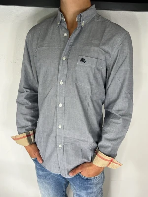 Burberry Skjorta  - Stilren grå skjorta med klassisk krage och vita knappar. Skjortan har en bröstficka med diskret logga och kontrasterande rutigt tyg på insidan av manschetterna. Perfekt till jeans för en avslappnad look.
